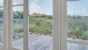 A view through a window with a wooden deck, leading to a beach with sand dunes and a house in the background.