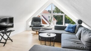 Een moderne loft slaapkamer met een schuine kap, een groot raam, een zwarte leren fauteuil, een grijze bank en een klein rond tafeltje.