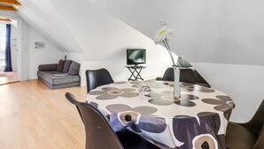 Een eethoek met een ronde tafel, bedekt met een tafelkleed met een patroon, omringd door stoelen. Een televisie is aan de muur bevestigd, en er staat een vaas met een bloem op de tafel.
