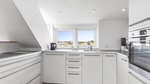 Een moderne keuken met witte kasten, een zwarte waterkoker en uitzicht op de omgeving door twee ramen.