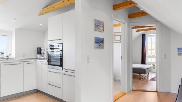Een moderne keuken met witte kasten, een ingebouwde oven en een koffiezetapparaat. Er is een houten plafond met zichtbare balken, en een uitzicht op een slaapkamer met een bed en een raam.