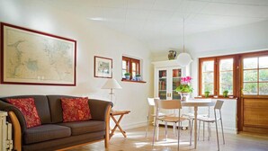 A cozy living room with a sofa, a dining table with chairs, a map on the wall, and a window with plants.