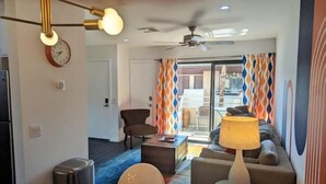 A modern living room with a sofa, armchair, and a glass coffee table. There is a ceiling fan, a lamp, and a clock on the wall. - PalmSpringsBNB - Renovated 1Bd with Amazing Views (Palm Springs)