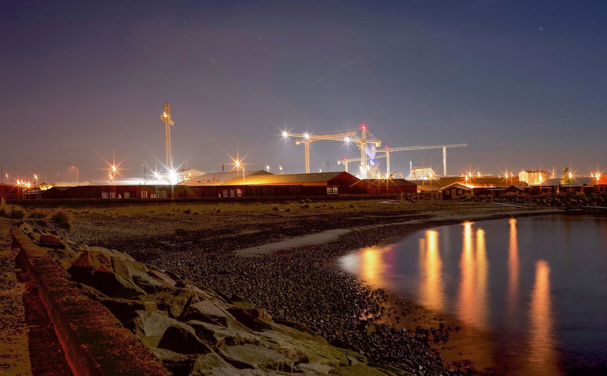 Area industriale notturna con gru ed edifici, una costa rocciosa e acque calme che riflettono le luci.