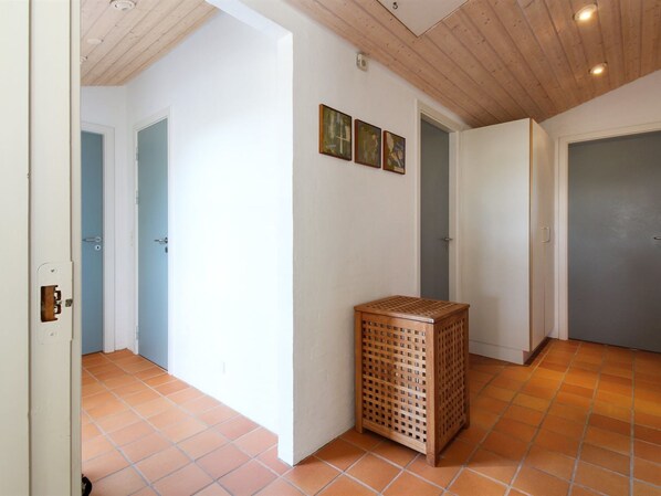A hallway with white walls, a wooden ceiling, and a tiled floor. There are three doors, one of which is open. A small wooden basket is placed against the wall.