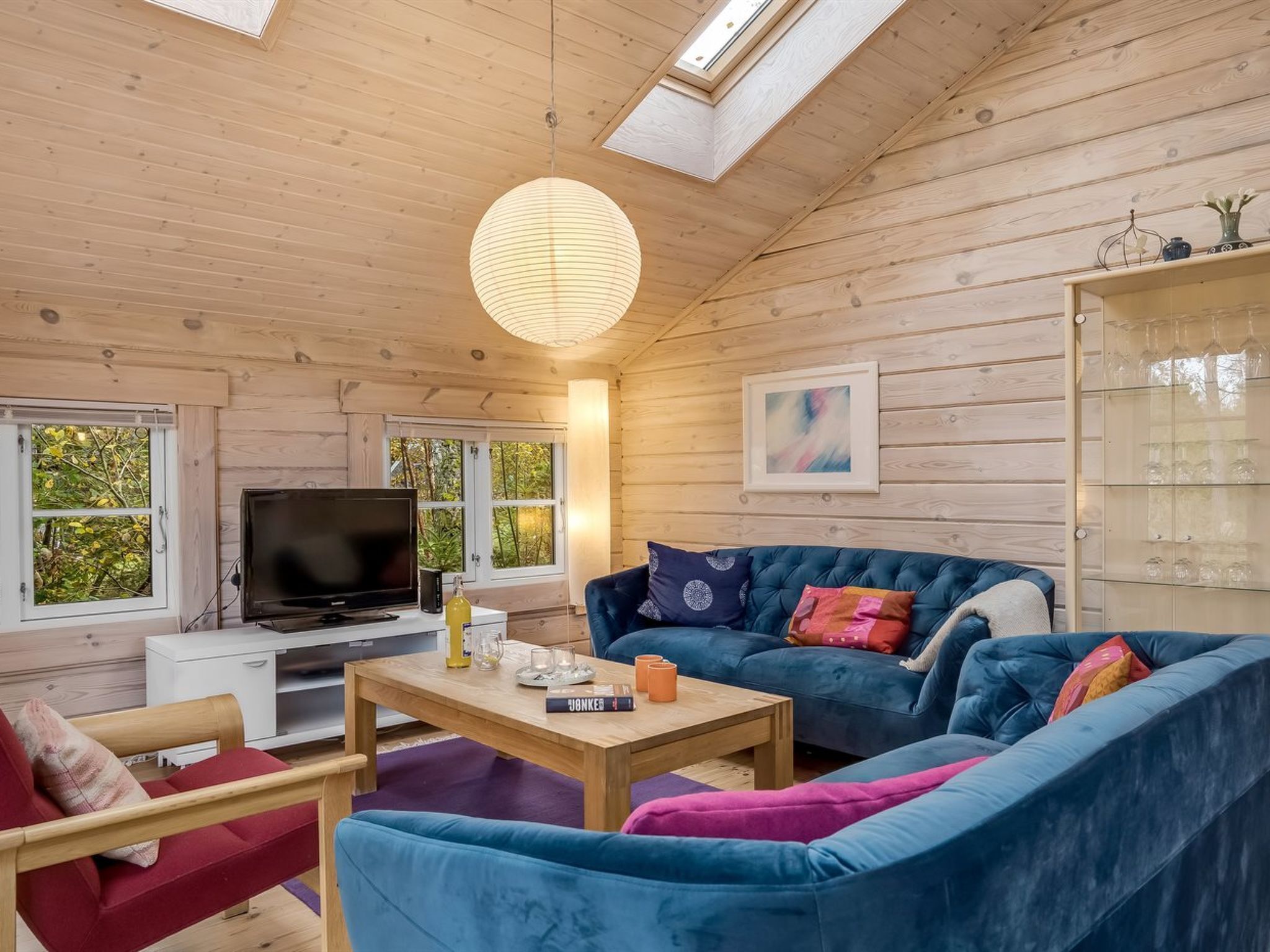 Un salon chaleureux avec un canapé bleu, une table basse en bois et un téléviseur. De grandes fenêtres offrent une vue sur la verdure environnante.