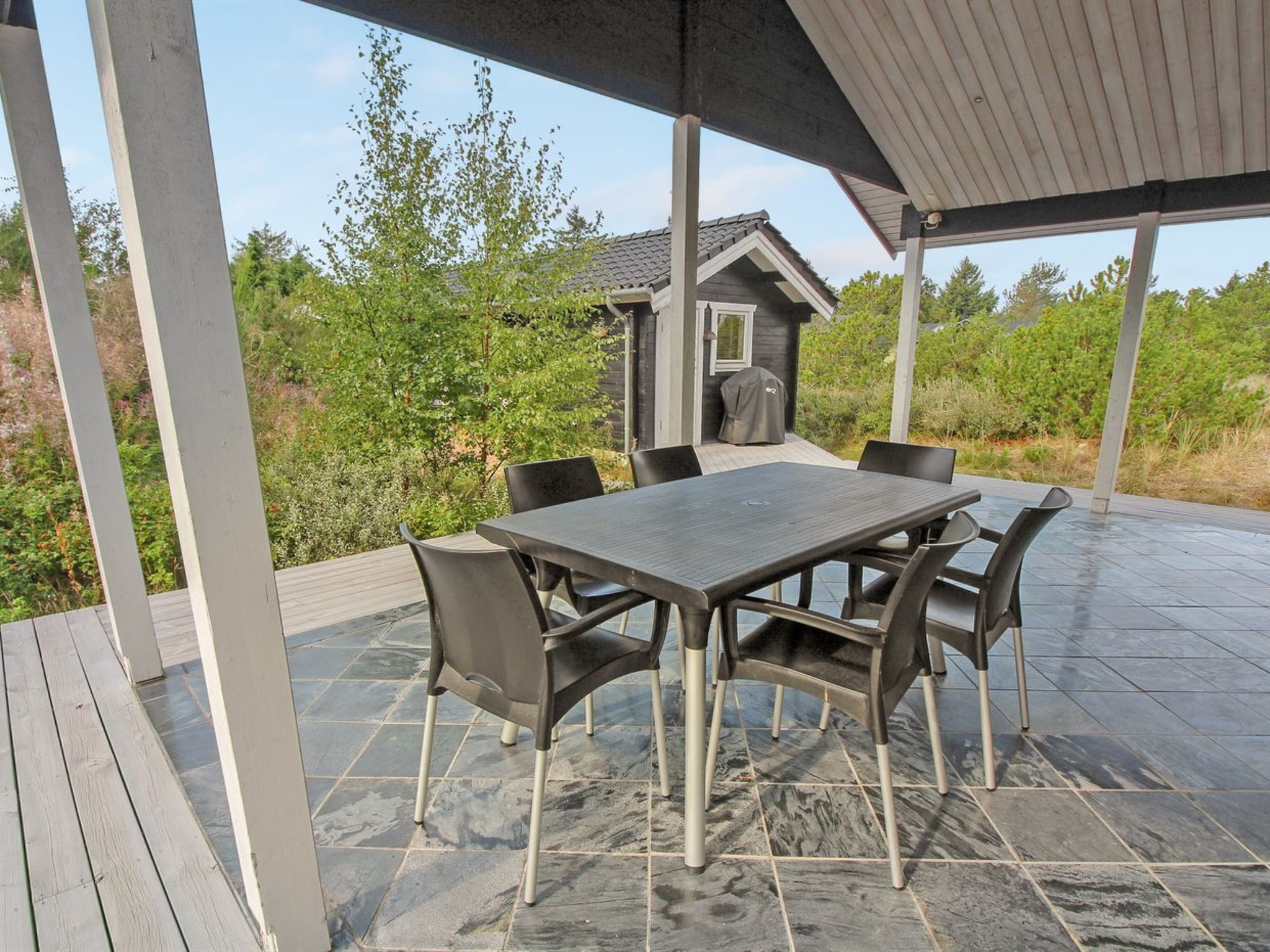 An outdoor patio with a stone-tiled floor, a rectangular table with chairs, and a covered area with a view of a house and greenery.