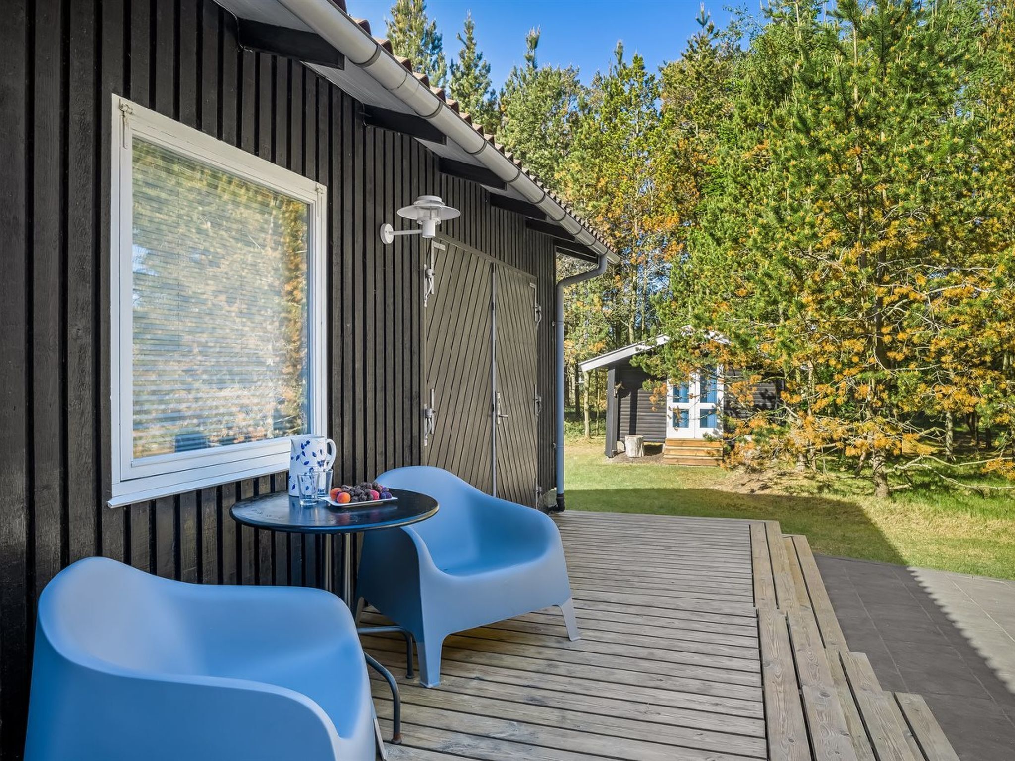 Een houten terras met een tafel, twee stoelen, een raam en een uitzicht op bomen en een gebouw op de achtergrond.