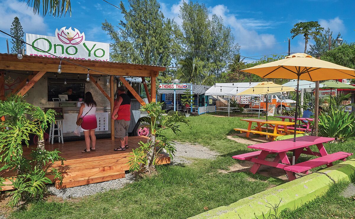 Un patio de comidas al aire libre con un letrero que dice 'Qno Yo', un puesto de comida tailandesa llamado 'Thongs Thai' y mesas de picnic.