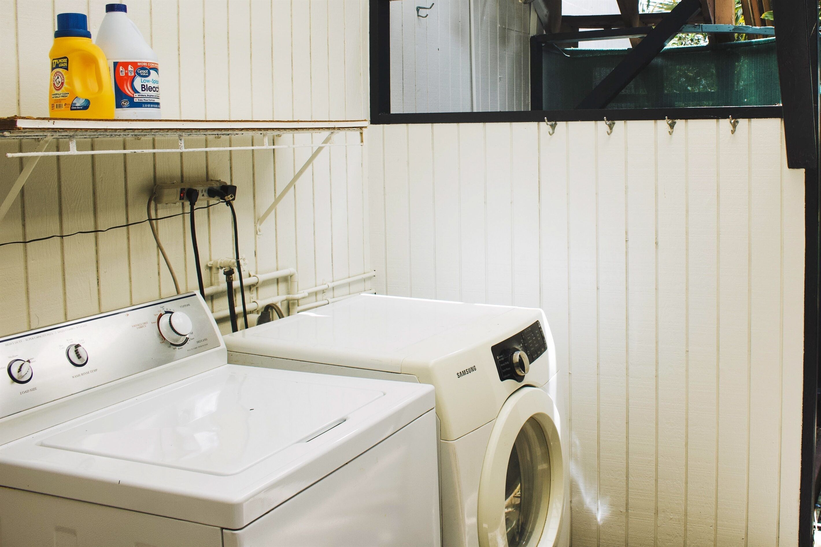 Un espace buanderie comprenant un lave-linge et un sèche-linge à chargement frontal, une étagère avec des produits de nettoyage et une fenêtre située au-dessus du sèche-linge.