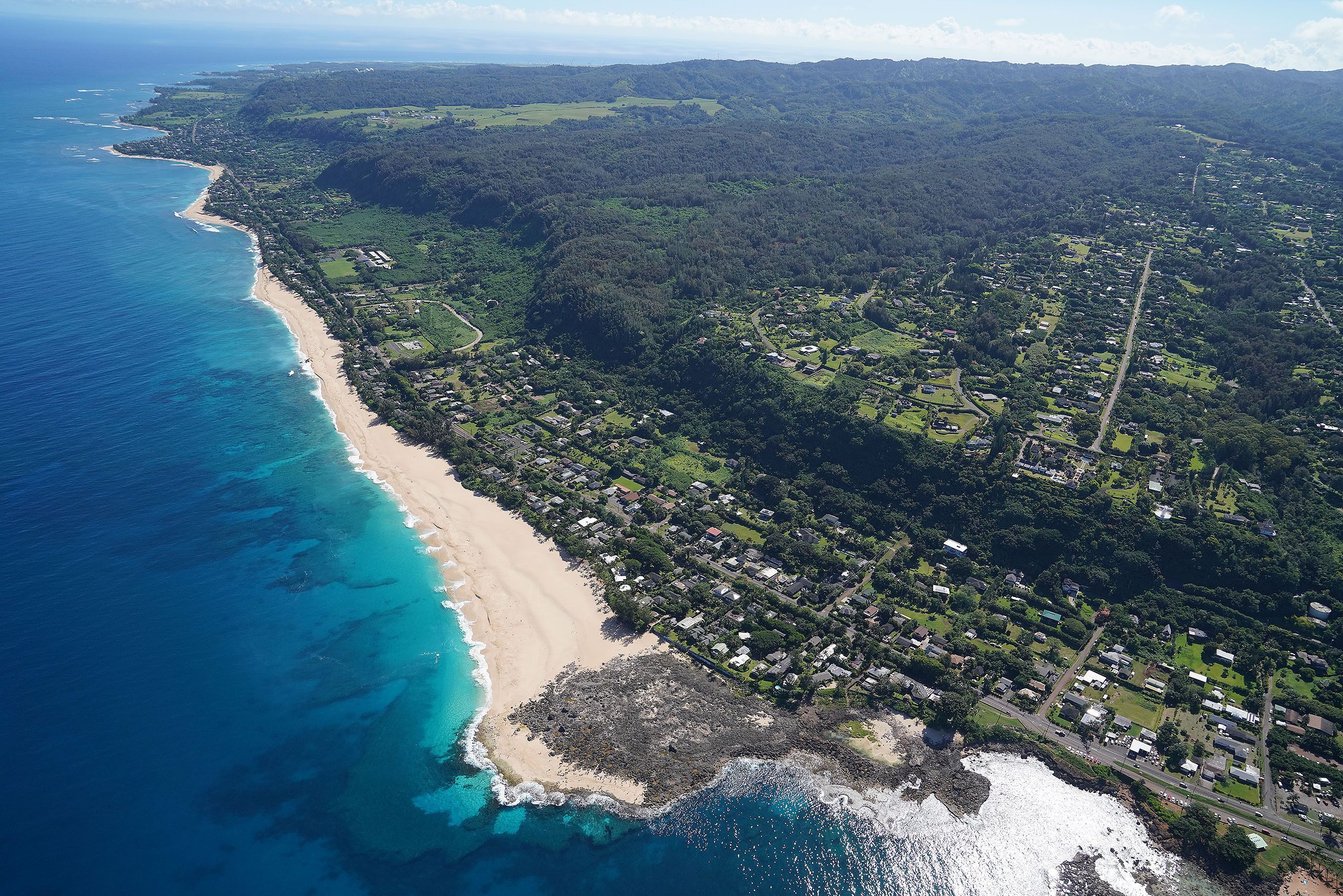 An aerial view of a coastal area with a beach, residential buildings, and surrounding greenery.