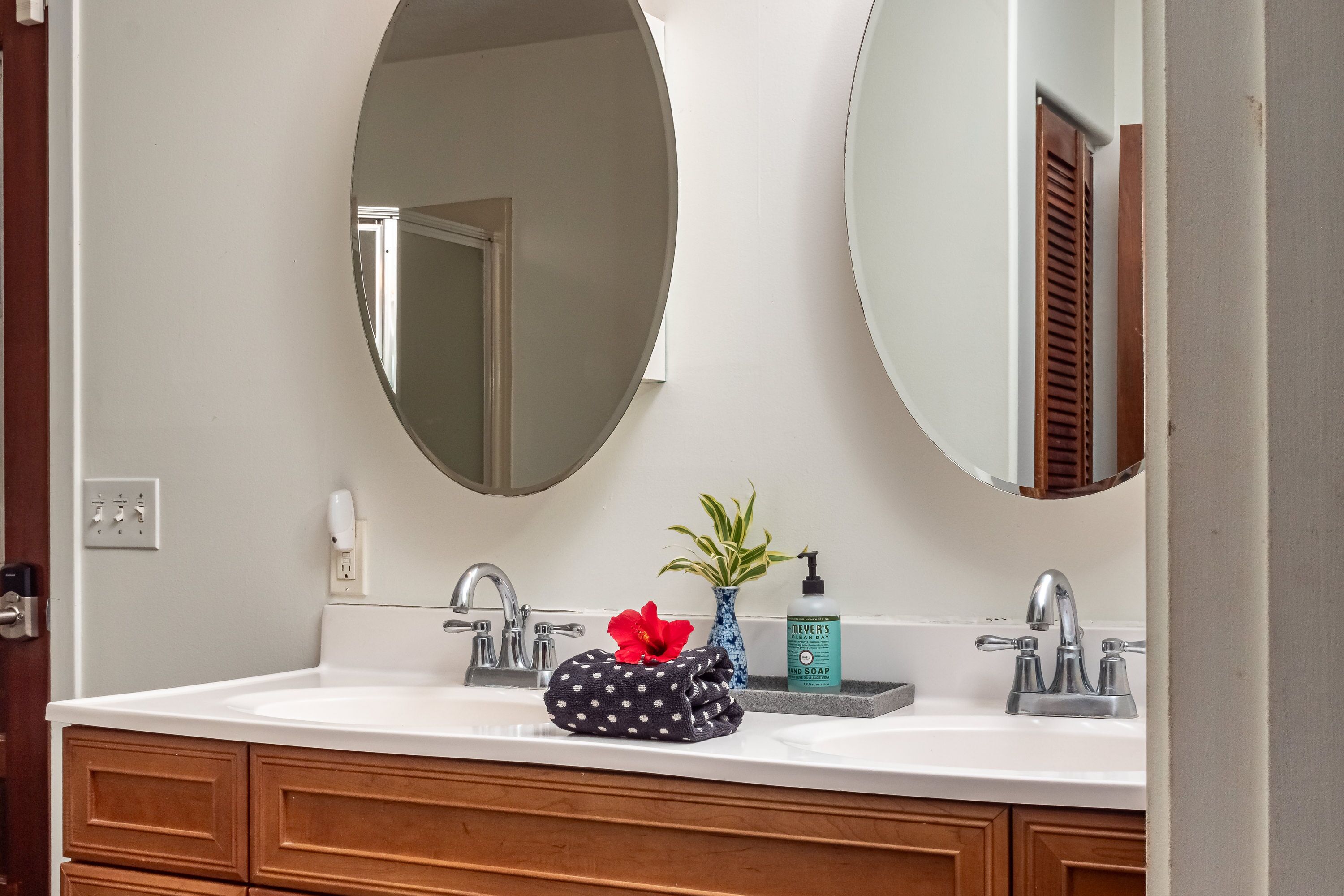 A bathroom with a white sink, two round mirrors, a wooden cabinet, a red flower, a blue vase with a plant, and a soap dispenser.