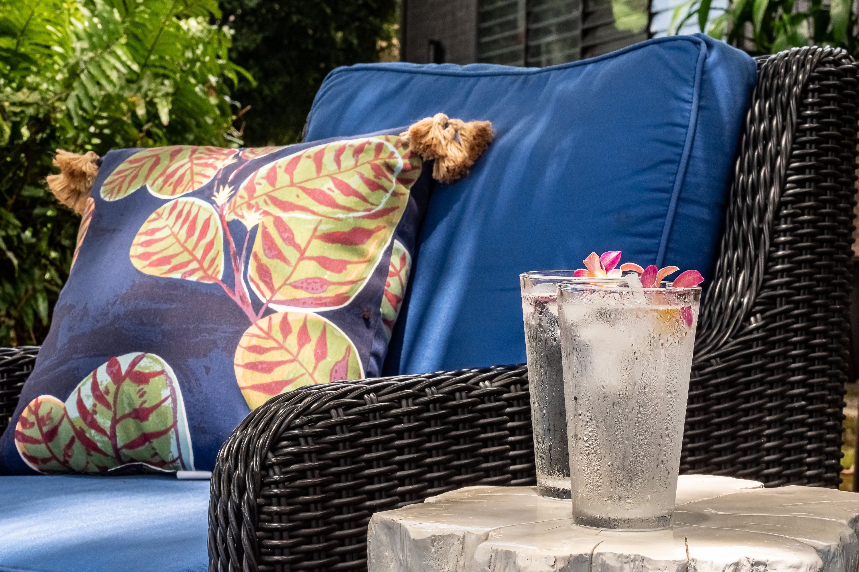 A wicker sofa with a blue cushion and patterned pillows, a clear glass with a drink, and a stone table.