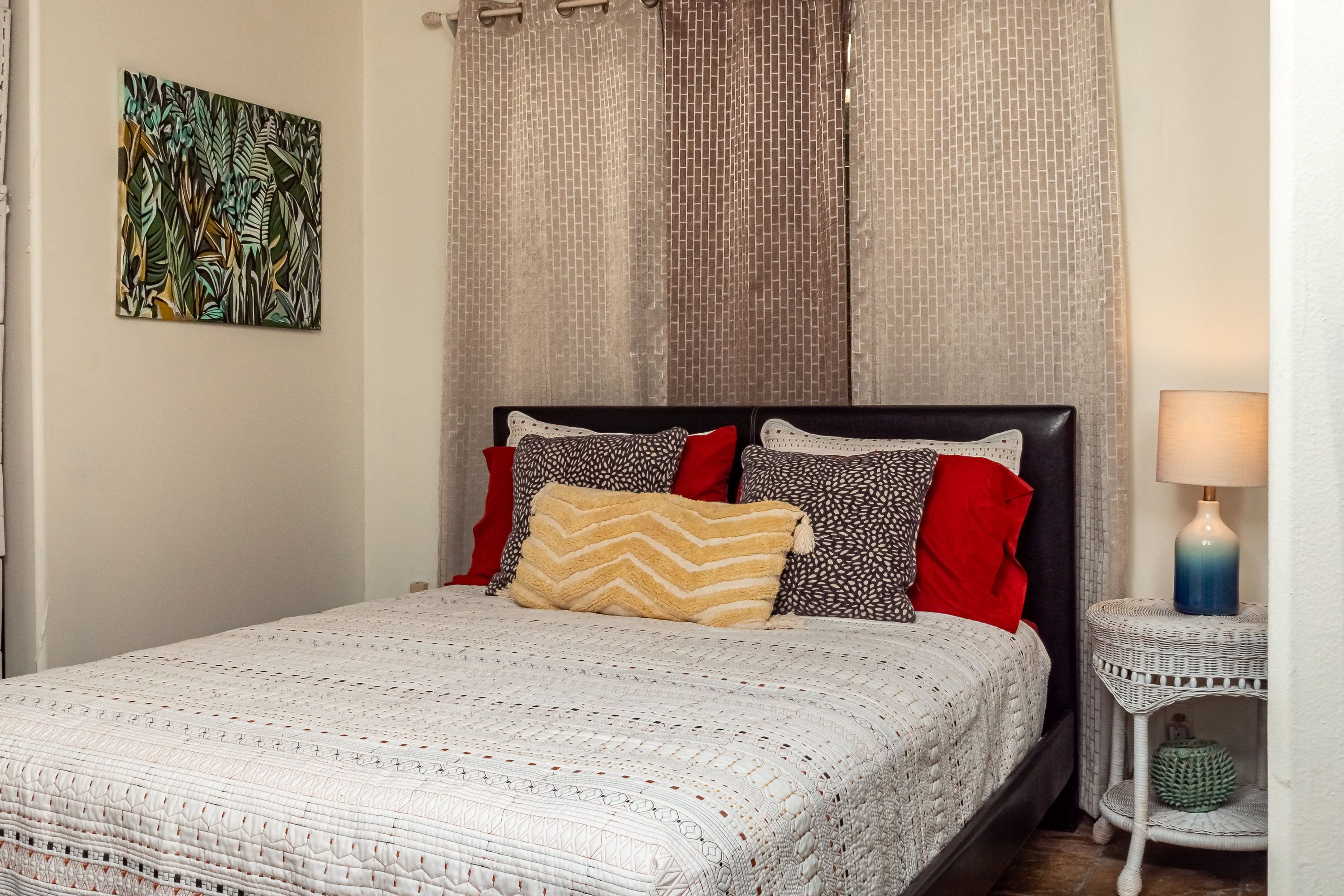 Una cama bien hecha con una colcha estampada y varias almohadas, un cuadro abstracto en la pared y una mesita de noche con una lámpara.