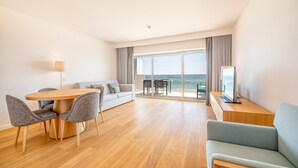 Habitación de hotel moderna con zona de comedor, sofá, televisión y balcón con vista al mar.