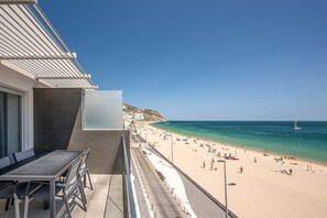 Ein Balkon mit Tisch und Stühlen mit Blick auf einen Strand, auf dem sich Personen und ein Segelboot befinden.