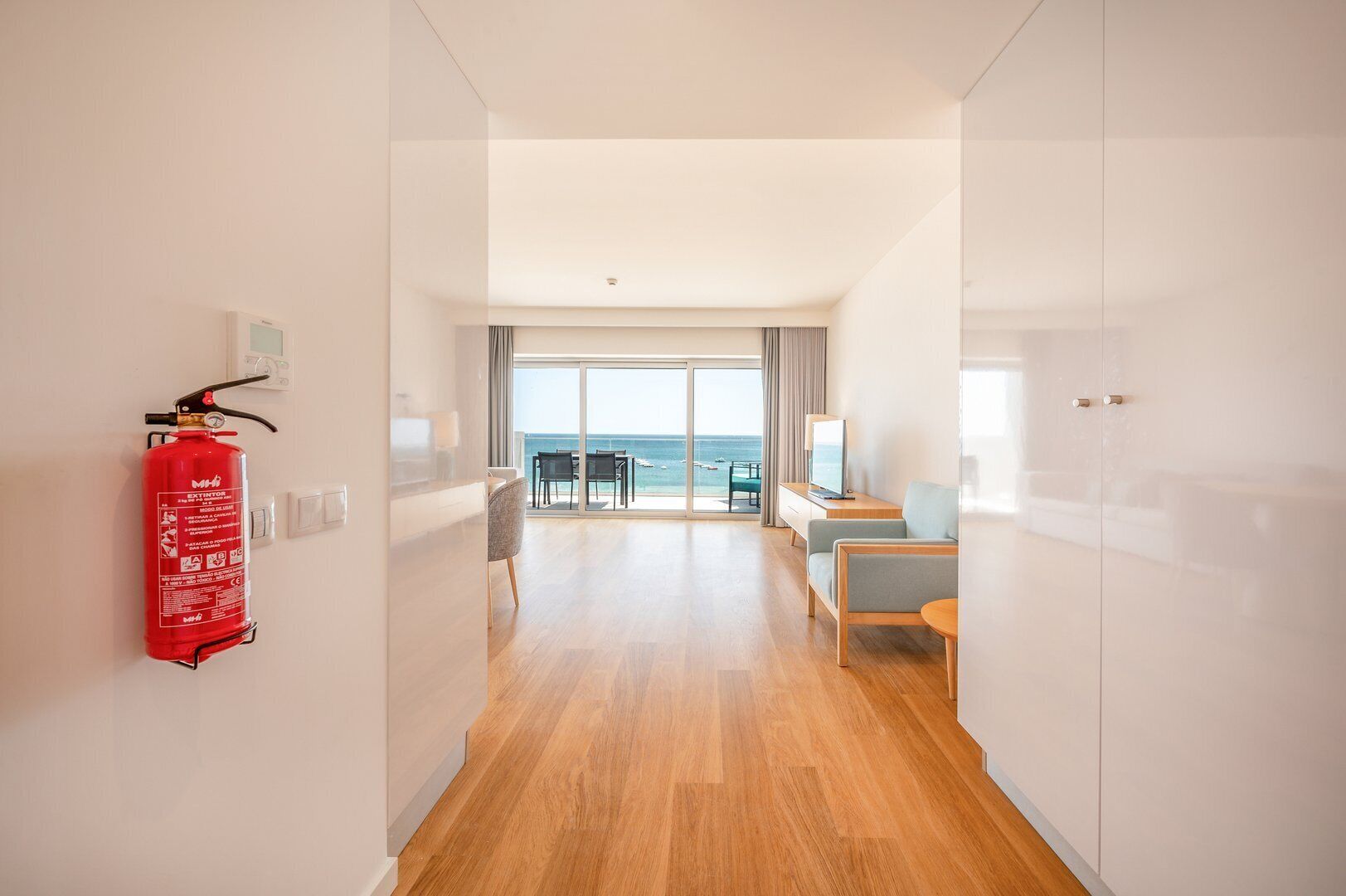 Pasillo de hotel con extintor, zona de comedor con vista al mar y área de estar con televisor.