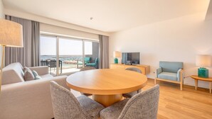 Habitación de hotel moderna con un balcón grande, una mesa de comedor redonda de madera, un sofá y un televisor sobre un mueble de madera.