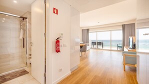 Una habitación de hotel moderna con cabina de ducha de vidrio, un extintor montado en la pared, un área de comedor con vista al mar y un balcón con mesa y sillas.