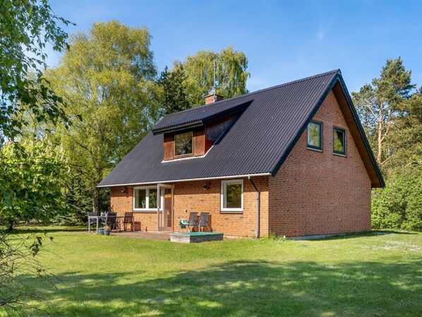 Une maison de plain-pied en briques, avec un toit en pente, une véranda à l’avant avec une table et des chaises, et une petite terrasse.