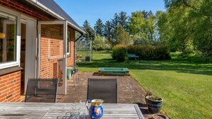 Une terrasse avec une table et des chaises, un mur en briques, une porte vitrée et un jardin agrémenté d’un banc et d’une serre.