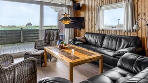 A cozy room with a wooden ceiling, a black leather sofa, wicker chairs, a wooden coffee table, and a large window offering a view of the countryside.