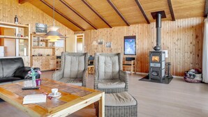 A cozy cabin interior with a wooden interior, a sofa, two armchairs, a table with a book and glasses, and a wood-burning stove.