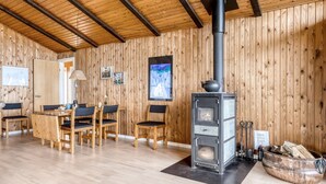 A cozy wooden interior with a central wood-burning stove, a dining area with chairs, and a television mounted on the wall.
