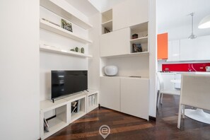 Un soggiorno moderno con una TV a schermo piatto su un mobile bianco, scaffali bianchi con oggetti decorativi e una parete rossa nell'area cucina.