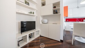 A modern living space with a flat-screen TV on a white console, white shelving with decorative items, and a red accent wall in the kitchen area. - Comfort Corner in Via Dante (Cagliari)