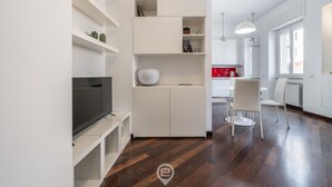 A modern living room with a flat-screen TV, white shelving, and a dining area with a red table and white chairs. - Comfort Corner in Via Dante (Cagliari)