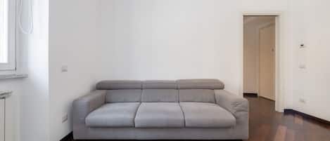 Un salón con tonos neutros, un sofá gris, suelo de madera y una pared blanca.