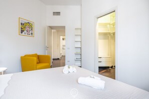 Una camera d'albergo con un letto, una poltrona gialla, una parete bianca con un quadro appeso e una cabina armadio.