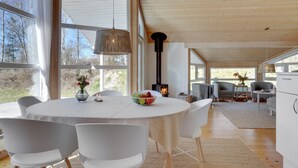 Een ruime kamer met een grote ronde tafel, witte stoelen, een moderne hanglamp en een houtkachel.