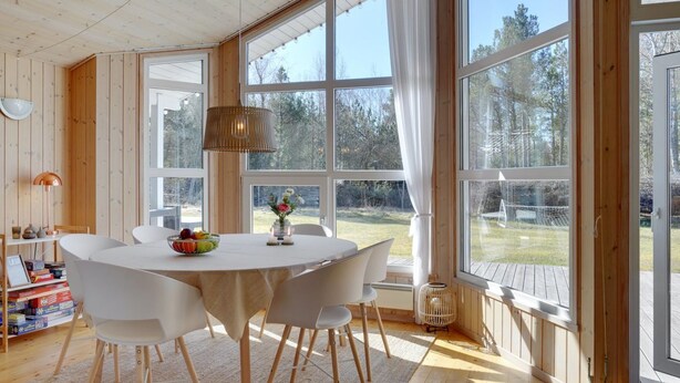 Een ruime kamer met een grote houten tafel, witte stoelen en uitzicht op een bos door grote ramen.