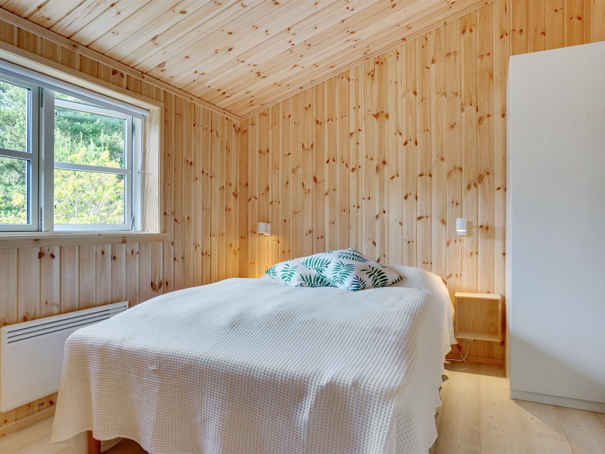 Ein Schlafzimmer mit Holzwandverkleidung, einem Bett mit weißer Bettdecke, zwei Nachttischlampen und einem Fenster mit Blick auf Grünflächen.