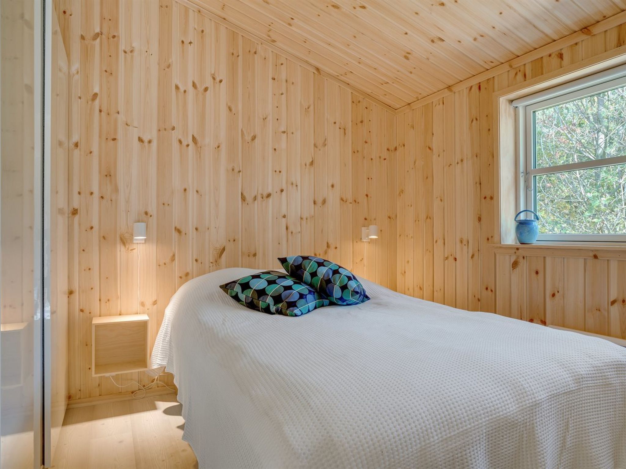 A room with wooden panel walls, a bed with white bedding and patterned pillows, a window with a view of trees, and two wall-mounted lights.