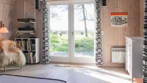 Une pièce chaleureuse aux murs en bois, avec une grande porte-fenêtre coulissante donnant sur une terrasse, un fauteuil moelleux, une bibliothèque et un meuble blanc.