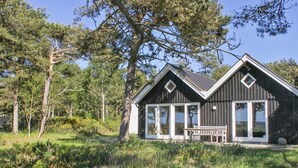 Une cabane en bois dotée de grandes fenêtres, d’un porche et entourée d’un paysage naturel.