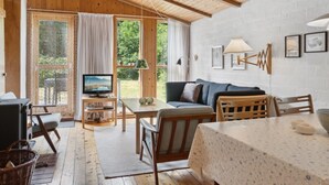Una camera accogliente con soffitto in legno, una TV su un mobile, un divano, un tavolo da pranzo e vista sul verde circostante.