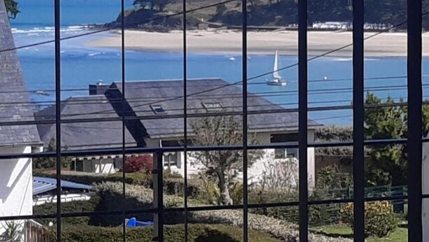 Udsigt over kysten gennem et vindue med et gittermønster, der viser en strand, hav og fjerne bakker.