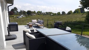 En udendørs terrasse med et bord med glasplade, fletmøbler og en grill.