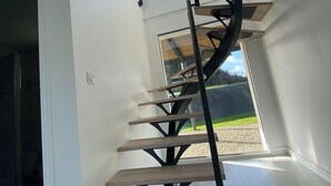 En moderne spiraltrappe med trætrapper og sorte metalgelændere, der fører op til et lyst rum med store vinduer og udsigt til det fri.