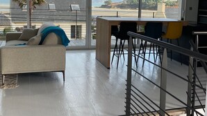 En moderne stue med sofa, spiseplads med stole, et køkken og store vinduer med udsigt til havet og bygninger.