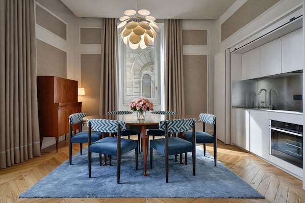 A dining area with a round table, six chairs, a chandelier, and a kitchen with modern appliances.