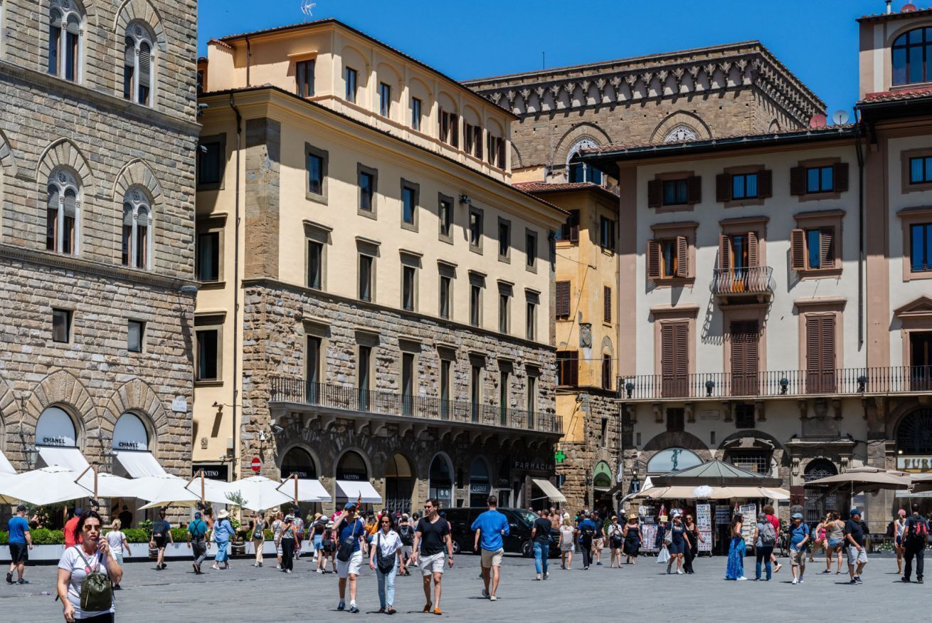 A historic square with stone buildings, outdoor cafes, and pedestrians.