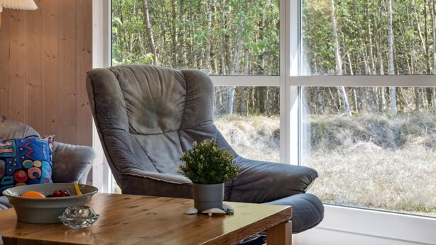 Ein Zimmer mit einem großen Fenster, einem grauen Sessel, einem kleinen Tisch mit einer Schale und einer Pflanze sowie einer Lampe links.
