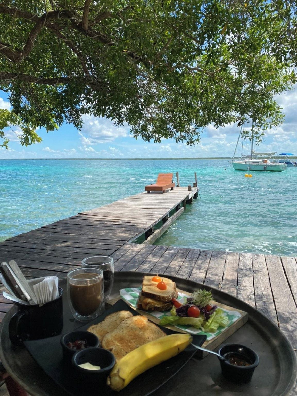 Una terraza de madera con una mesa dispuesta para una comida, con vista a un mar azul y transparente con barcos a lo lejos.
