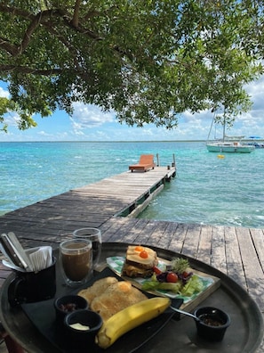 A wooden deck with a table set for a meal, overlooking a clear blue sea with boats in the distance. - Glamping Asana Bacalar Breakfast & Kayak (Bacalar)