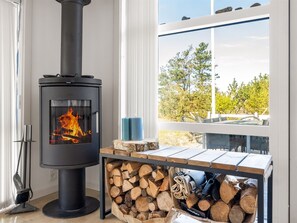 A modern room with a wood-burning stove, a window with a view of trees, and a stack of firewood.